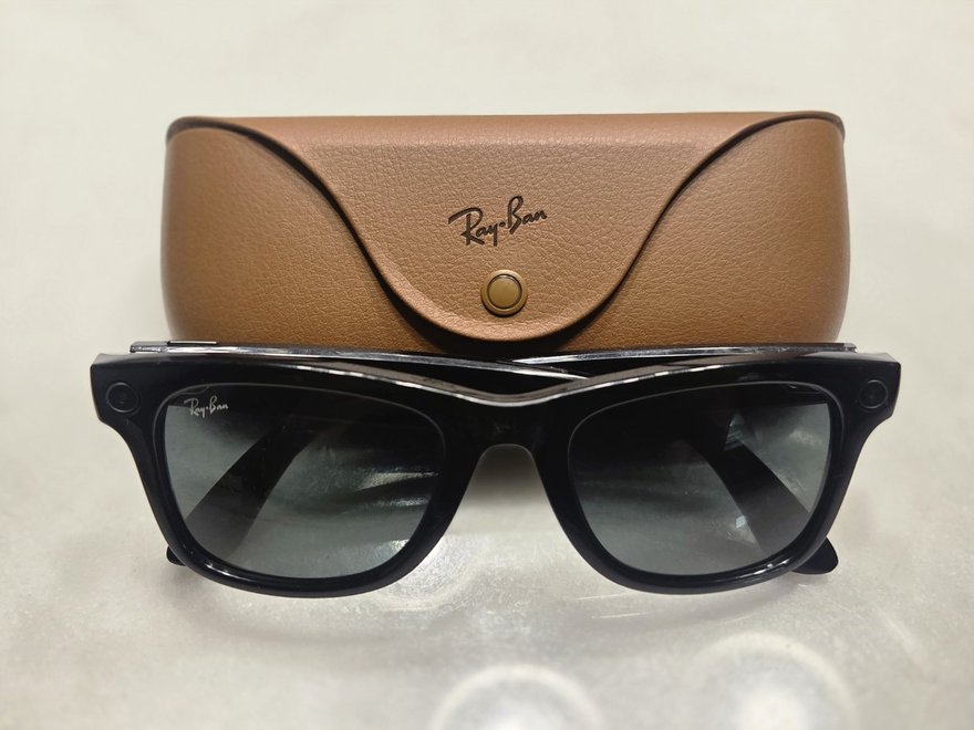 Kahverengi deri şarj kutusunun önünde siyah çerçeveli Ray-Ban Meta gözlük; çerçevenin köşelerinde küçük kamera ve ışık
