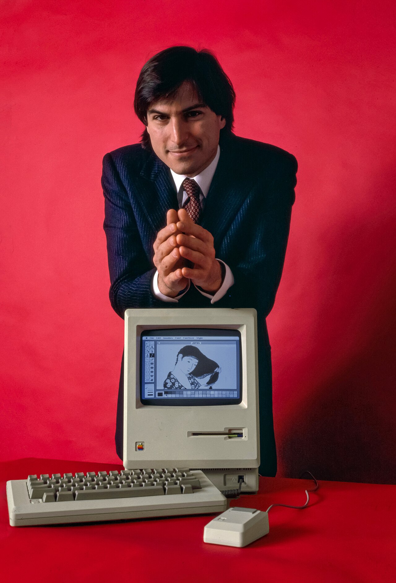 Kırmızı fon önünde genç Steve Jobs ve küçük bej bir Macintosh bilgisayar