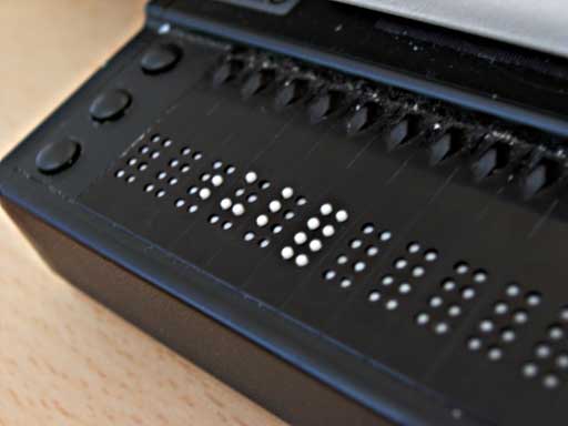 Bir dizüstü bilgisayarın klavyesinin önünde duran yenilenebilir Braille ekranı: üzerinde iğnelerin yükselip alçalarak harfleri oluşturduğu uzun bir şerit.